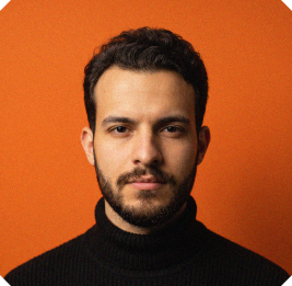 Portrait of a bearded man with dark hair wearing a black turtleneck, looking directly at the camera against a solid orange background.